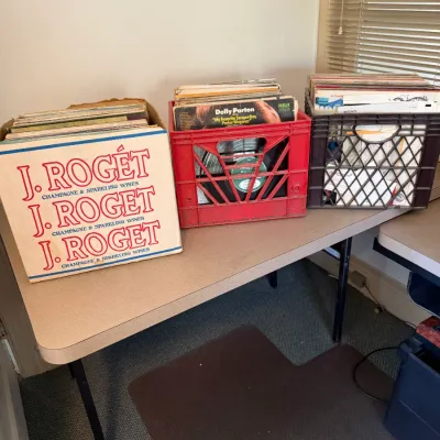 Sale Photo Thumbnail #188: Collection of vinyl LP records stored in a J. Rogét cardboard box and two plastic crates, displayed on a folding table.