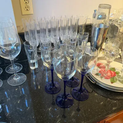 Sale Photo Thumbnail #157: Collection of glassware featuring wine glasses with cobalt blue stems, Ramazzotti branded shot glasses, champagne flutes, and a stainless steel cocktail shaker on a black granite countertop.