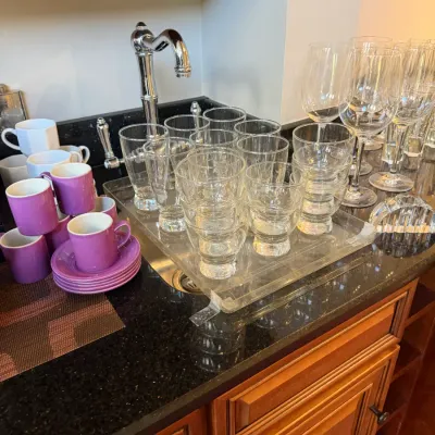Sale Photo Thumbnail #155: Collection of glassware and ceramic coffee mugs arranged on a wet bar countertop, including clear tumbler sets, wine glasses, and purple espresso cups with matching saucers.
