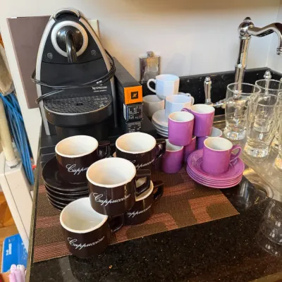 Sale Photo Thumbnail #154: Nespresso Essenza espresso machine displayed on a countertop alongside coffee pods, sets of brown cappuccino cups, and purple espresso cups with saucers.