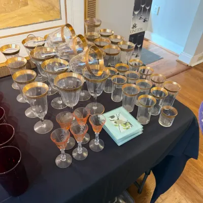 Sale Photo Thumbnail #162: Large collection of gold-rimmed crystal glassware including stemmed wine glasses, highball tumblers, and liqueur glasses, displayed alongside a stack of decorative coasters.
