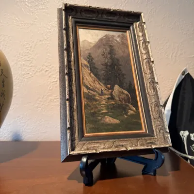 Sale Photo Thumbnail #94: Framed miniature landscape oil painting on a tabletop easel, depicting a mountainous scene in a carved silver and black frame.