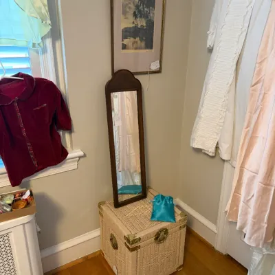 Sale Photo Thumbnail #131: Painted white wicker trunk with brass hardware, featuring a wooden wall mirror resting on top and framed landscape art above.