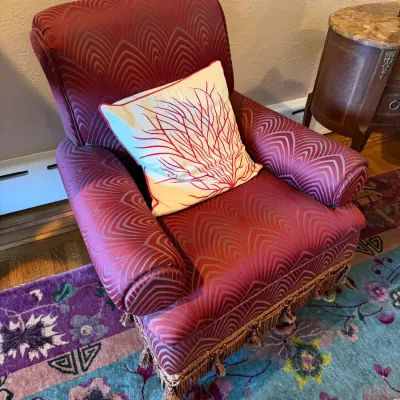 Sale Photo Thumbnail #96: Red patterned armchair featuring rolled arms, fringe trim along the bottom, and clear casters, styled with a coral-embroidered throw pillow.