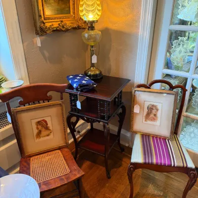 Sale Photo Thumbnail #90: Vintage interior scene featuring framed portrait art resting on wooden chairs, an illuminated yellow glass oil-style lamp on a spool-leg side table, and an ornate gold framed painting.