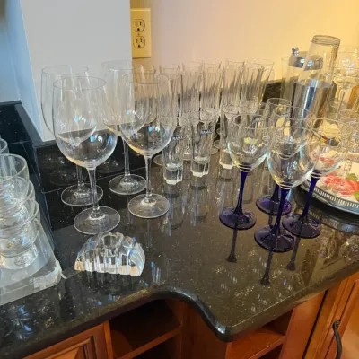 Sale Photo Thumbnail #156: Large assortment of glassware, including wine glasses, cobalt blue-stemmed glasses, champagne flutes, shot glasses, and a stainless steel cocktail shaker set arranged on a dark countertop.