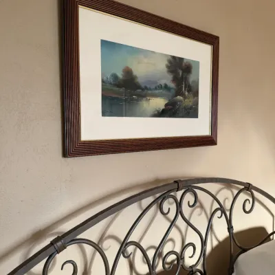 Sale Photo Thumbnail #142: Framed landscape art print featuring a tranquil lake scene, presented in a dark wooden ribbed frame with a gold trim inner detail, positioned above an arched wrought iron headboard.