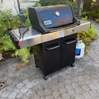 Sale Photo Thumbnail #226: Weber Spirit propane gas grill with stainless steel side shelves, black double-door storage cabinet, and a attached propane tank.