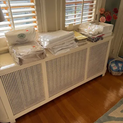 Sale Photo Thumbnail #169: Selection of folded bed linens and sheet sets in original packaging displayed atop a long white lattice radiator cover.