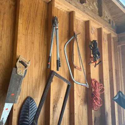 Sale Photo Thumbnail #219: Assortment of garage and garden tools mounted on a wooden shed wall, including hand saws, loppers, extension cords, and decorative frame elements.