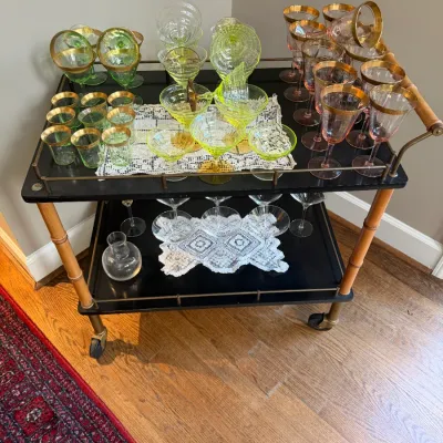 Sale Photo Thumbnail #153: Mid-century faux bamboo black rolling bar cart displayed with various vintage glassware collections, including gold-rimmed stems and vaseline or depression glass pieces.