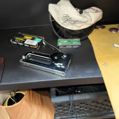 Sale Photo Thumbnail #152: Chrome staple gun alongside boxes of Arrow staples and a white printed visor cap on a black shelf.