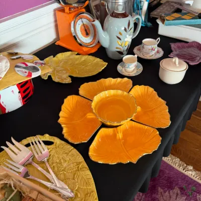 Sale Photo Thumbnail #108: Collection of vintage kitchenware and serving items displayed on a table, including an orange leaf-shaped chip and dip set, a mid-century orange kitchen scale, a hand-painted coffee pot, and silver-plated serving forks.