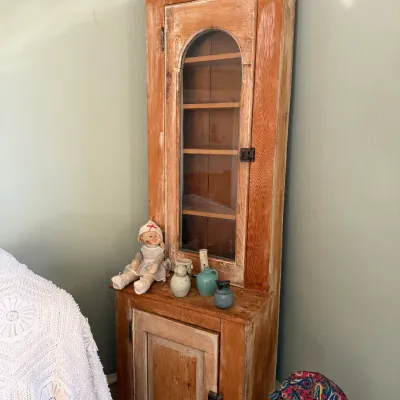 Sale Photo Thumbnail #137: Rustic tall pine corner cabinet with an arched glass door over a lower wooden cupboard, styled with vintage ceramic pieces and a cloth doll.