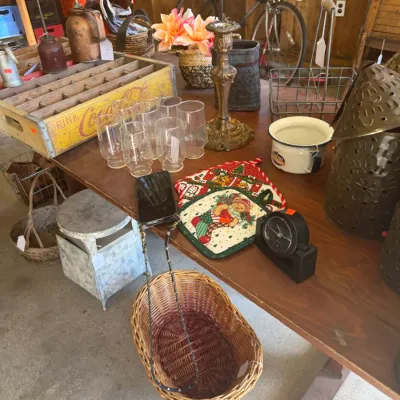 Sale Photo Thumbnail #212: An assortment of vintage housewares and collectibles displayed on a wooden table, including a yellow Coca-Cola crate, drinking glasses, a brass candlestick, and a woven basket.