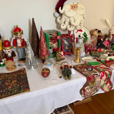 Sale Photo Thumbnail #179: Collection of Christmas decorations displayed on a table, including nutcrackers, stockings, tabletop trees, a large plush Santa face, and boxed ornament sets.