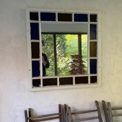 Sale Photo Thumbnail #79: Square wall mirror framed in a white painted multi-pane window with blue and amber stained glass inserts, mounted above vintage wooden folding chairs.