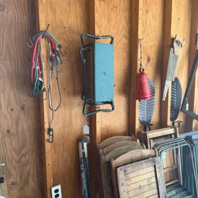Sale Photo Thumbnail #217: Collection of garage items including folding wooden and metal chairs, bungee cords hanging on a wall, a hand saw, and decorative wall hangings.
