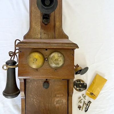 Antique oak wall-mounted hand-crank telephone featuring brass ringer bells, side receiver, and various small loose replacement parts.