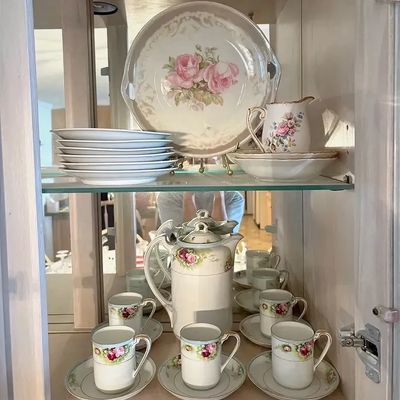 Vintage floral porcelain tea and dessert set displayed in a glass-shelf cabinet, including a tall coffee pot, demitasse cups with...