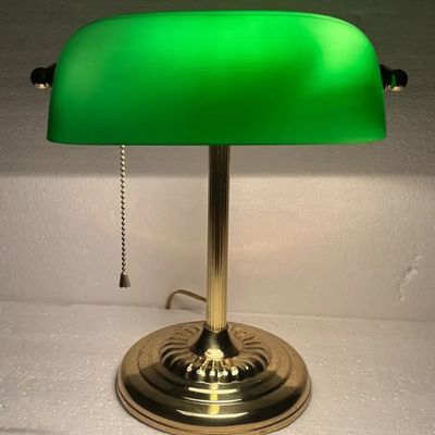 Classic banker's desk lamp featuring a polished brass base, fluted column stem, bead-chain pull switch, and an illuminated green...