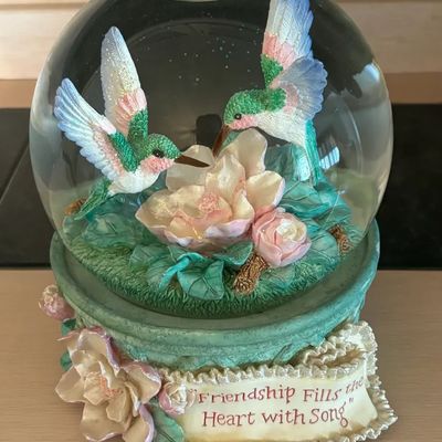 Decorative hummingbird snow globe featuring two iridescent hummingbirds hovering over pink blossoms, sitting on an ornate base inscribed...
