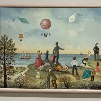Framed folk art style oil painting depicting figures flying kites and hot air balloons along a coastline, presented in a light wooden frame.