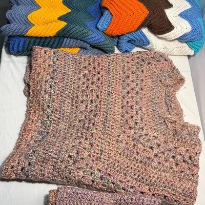 Collection of handmade crochet items, including colorful ripple-patterned chevron afghans and muted pink variegated yarn blankets.