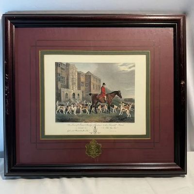 Framed English equestrian hunt print depicting a huntsman on horseback surrounded by hounds in front of an estate, set in a double-matted...