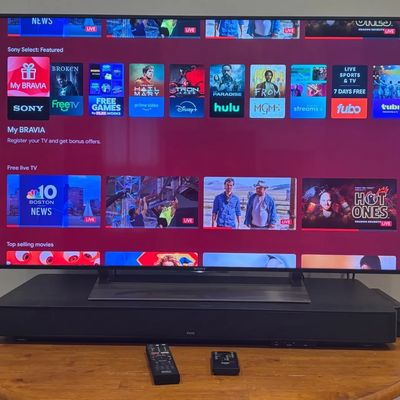 Sony Bravia flat-screen smart TV powered on, displayed on a wooden stand above a ZVOX soundbar with accompanying remotes.