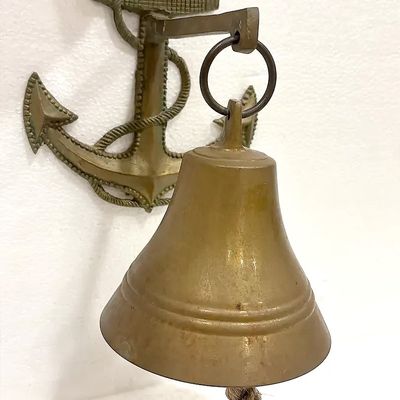 Vintage brass nautical ship's bell mounted on an anchor-shaped wall bracket, featuring a braided rope clapper pull.