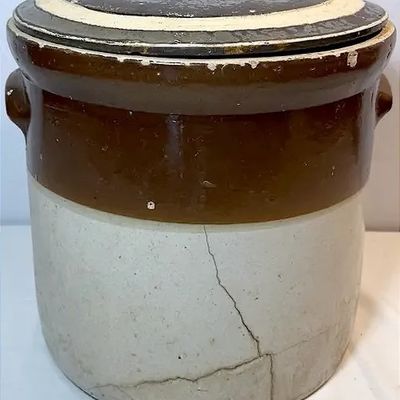 Antique two-tone stoneware crock with lid, featuring molded side handles and visible cracking along the lower body.
