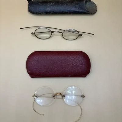 Two pairs of antique eyeglasses with matching protective cases, including small oval-framed spectacles and rimless wire-rimmed glasses...