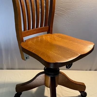 Vintage oak banker's swivel desk chair featuring a slat back design, contoured seat, and caster wheels.