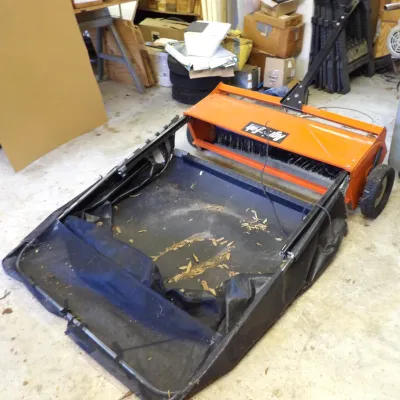 Agri-Fab tow-behind lawn sweeper featuring an orange steel housing, rotating brush, tow bar, and large black mesh collection bag.