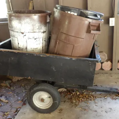 Black metal tow-behind lawn cart carrying two large brown plastic trash cans, showing visible wear and scuffs from heavy use.