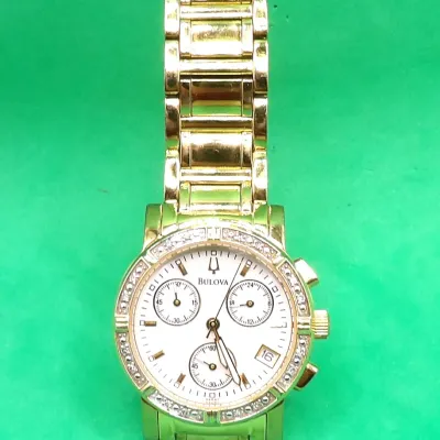 Bulova gold-tone quartz chronograph wristwatch featuring a crystal-accented bezel, white dial with three subdials, date window, and a...