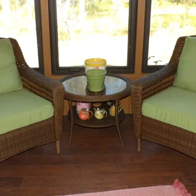 Pair of outdoor woven wicker armchairs with light green cushions and a matching round glass-top side table in a sunroom setting.