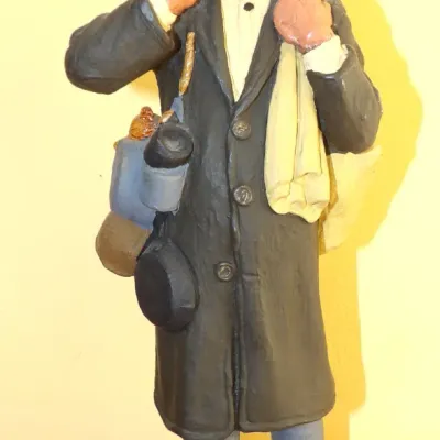 Hand-painted molded figurine of a peddler or traveling vendor, wearing a derby hat and coat while carrying wares including a frying pan...