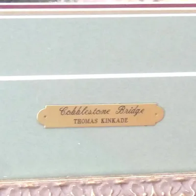Engraved brass title plate for Thomas Kinkade's 'Cobblestone Bridge,' set against a sage green double mat inside an ornate...