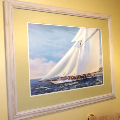 Framed nautical art print depicting a sailboat on open waters, presented in a light whitewashed wood frame with a wide coordinating mat...