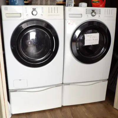 Matching Kenmore front-loading washing machine and electric dryer pair, finished in white with dark glass doors and mounted on storage...