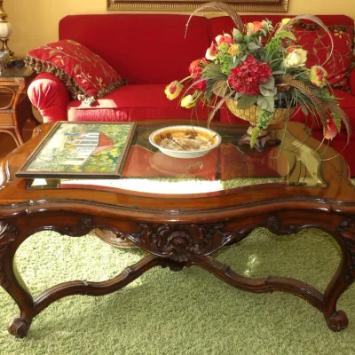 Ornate French Provincial style carved wood coffee table with a glass insert top, displaying a framed artwork, a shallow bowl, and an...