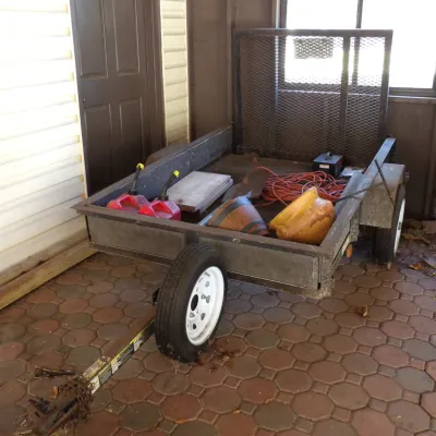 Single-axle utility trailer with a mesh fold-down rear ramp, loaded with assorted items including red gas cans, extension cords,...