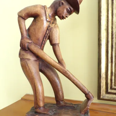 Hand-carved wooden sculpture depicting a barefoot worker wearing a hat and holding a hoe, featuring detailed craftsmanship on a carved base.