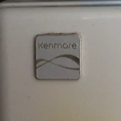 Close-up detail view of a metallic Kenmore brand logo emblem on a white household appliance.