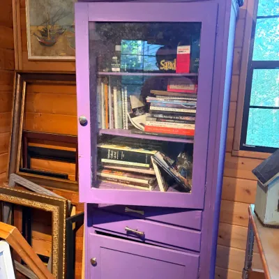 Sale Photo Thumbnail #138: Bright purple painted wooden display cabinet featuring a glass upper door filled with art books, two middle drawers, and a lower storage cupboard.