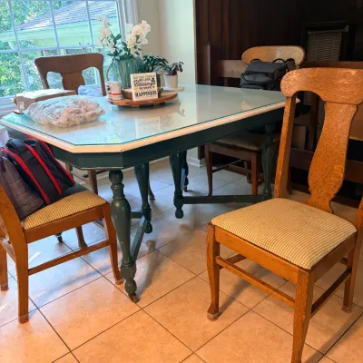 Vintage painted dining table with a glass top and turned legs, surrounded by matching oak side chairs with upholstered seats.