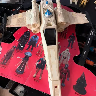 Sale Photo Thumbnail #108: Vintage Kenner Star Wars X-Wing Fighter toy vehicle from around 1978, resting atop a torn vintage character checklist insert.