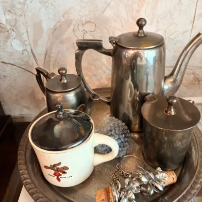 Vintage pewter or tarnished silver-tone tea and coffee set resting on a round tray, accompanied by decorative cork stoppers and a...